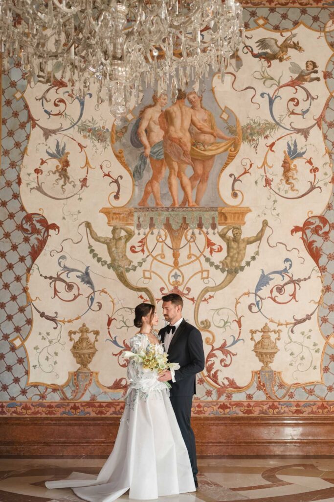 Hochzeit im Belvedere Wien: Brautpaar im Festsaal vor der bemalten Wand, Kristallluster über ihnen