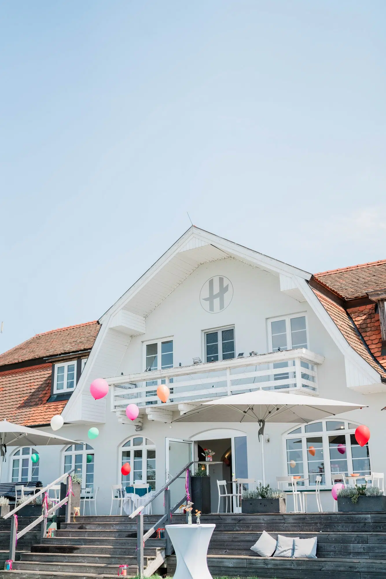 Weißes Giebelgebäude des Hannersberg mit bunten Luftballons und Sonnenschirmen vor blauem Himmel